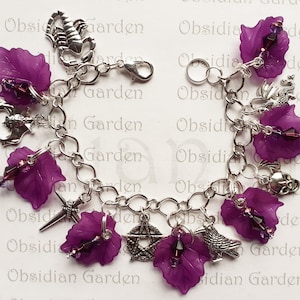 May include: A silver charm bracelet with purple flower charms, a silver bat charm, a silver scorpion charm, a silver pentagram charm, a silver cross charm, a silver bird charm, and a silver skull charm.