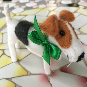 May include: A white and black felt toy dog with brown ears and a green bow. The dog is standing on a tiled surface.