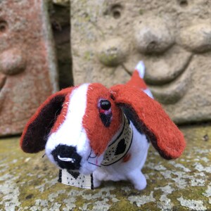 May include: A small, hand-crafted felt toy dog with brown, white, and black fur. The dog is wearing a black and white collar with a heart design.