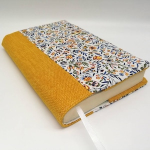 Könnte beinhalten: Ein gelber und weißer Buchumschlag aus Blumenstoff mit einem gelben Leinenrücken. Der Umschlag hat ein weißes Band als Lesezeichen.