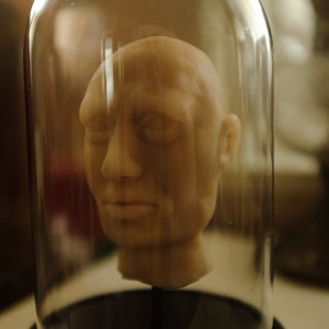 Könnte beinhalten: Eine Tonskulptur eines menschlichen Kopfes mit geschlossenen Augen wird unter einer Glasglocke auf einem schwarzen Sockel ausgestellt.