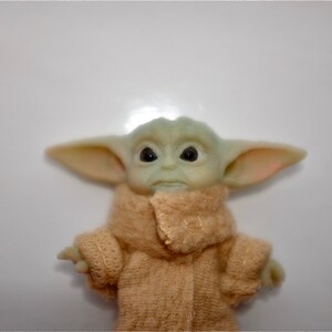 May include: A small green plush toy with large ears and big eyes, wearing a brown knitted coat. The toy is a popular character from the Star Wars franchise.