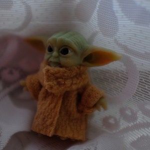May include: A small green and brown plush toy wearing a brown coat. The toy is sitting on a white lace fabric.