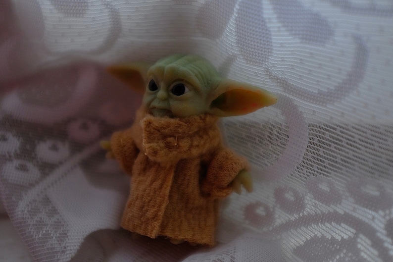May include: A small green and brown plush toy wearing a brown coat. The toy is sitting on a white lace fabric.