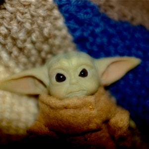 May include: A small green plush toy with large ears and big eyes, wearing a brown hooded robe. The toy is sitting on a blue and white fuzzy surface.