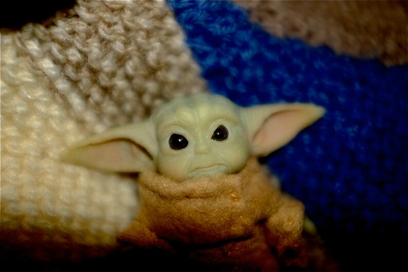 May include: A small green plush toy with large ears and big eyes, wearing a brown hooded robe. The toy is sitting on a blue and white fuzzy surface.