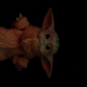 May include: A small, green, and brown plush toy with large ears and big eyes. The toy is wearing a brown knitted sweater.