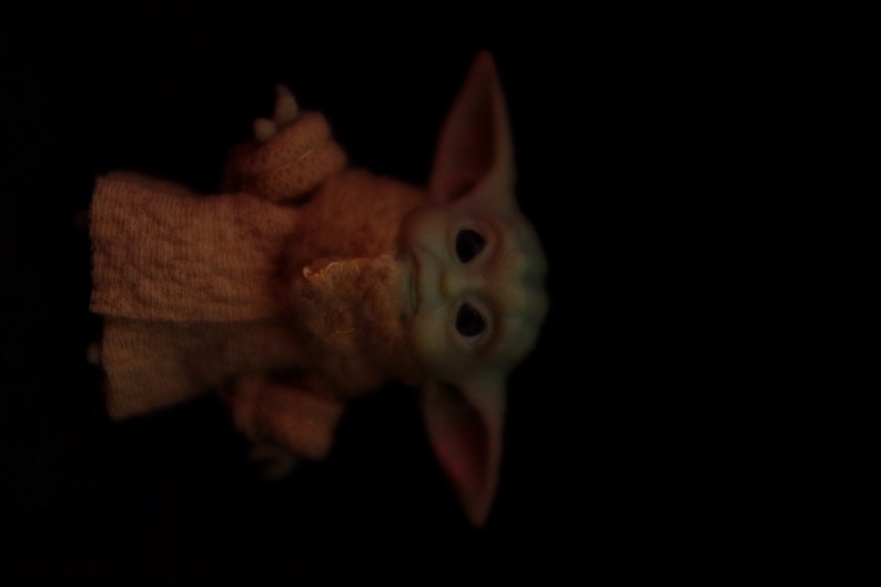 May include: A small, green, and brown plush toy with large ears and big eyes. The toy is wearing a brown knitted sweater.