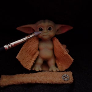 May include: A small, green, and brown figurine of a character from the Star Wars franchise. The figurine is wearing a brown, knitted jacket and has large ears. A paintbrush is being used to paint the figurine's face.