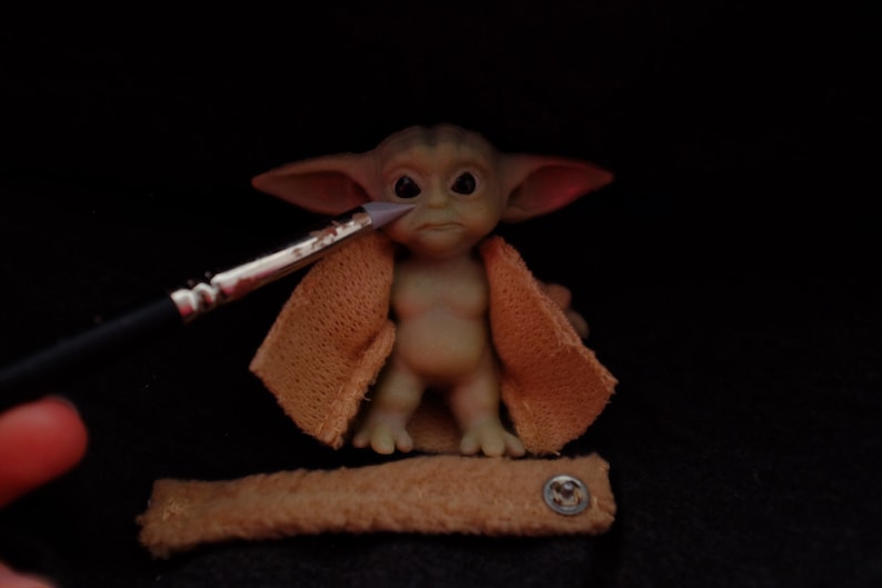 May include: A small, green, and brown figurine of a character from the Star Wars franchise. The figurine is wearing a brown, knitted jacket and has large ears. A paintbrush is being used to paint the figurine's face.