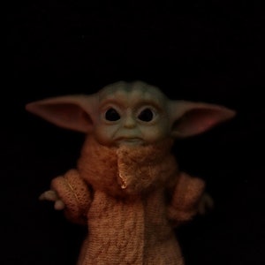 May include: A small, green, plush toy with large ears and big eyes wearing a brown, fuzzy coat. The toy is a popular character from the Star Wars franchise.