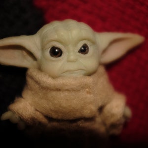 May include: A small green plush toy with large ears and big brown eyes. The toy is wearing a brown robe and has a serious expression.