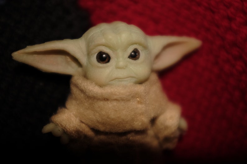 May include: A small green plush toy with large ears and big brown eyes. The toy is wearing a brown robe and has a serious expression.