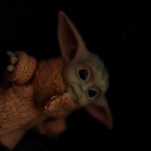 May include: A small, green, plush toy with large ears and big eyes. The toy is wearing a brown knitted sweater.