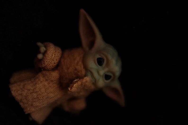 May include: A small, green, plush toy with large ears and big eyes. The toy is wearing a brown knitted sweater.