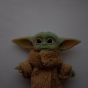 May include: A green and brown plush toy with large ears and big eyes. The toy is wearing a brown robe.