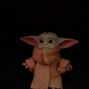 May include: A small green and brown plush toy with large ears and big eyes wearing a knitted orange coat.