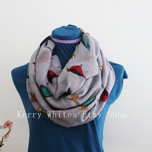 May include: A gray infinity scarf with a colorful bird print. The scarf is displayed on a blue mannequin.