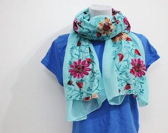 May include: A light blue scarf with embroidered floral designs in shades of pink, red, and orange. The scarf is draped over a blue shirt on a mannequin. The floral embroidery is detailed and adds a decorative element to the scarf.