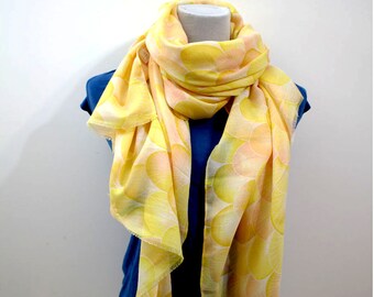 May include: A lightweight scarf in shades of yellow and peach, patterned with overlapping semi-circles. The scarf is draped over a blue top on a mannequin, showcasing its flowing fabric and design.