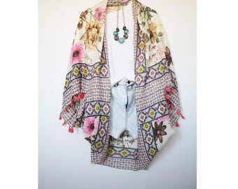 May include: A white floral kimono with pink and blue accents and tassels. The kimono is hanging on a hanger and has a white background.