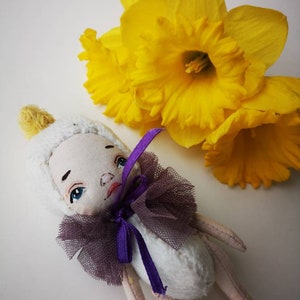Può includere: Una bambola di peluche bianca con un nastro viola e un colletto di tulle. La bambola ha un viso dipinto con occhi blu e una bocca rosa. La bambola è sdraiata su una superficie bianca con narcisi gialli sullo sfondo.