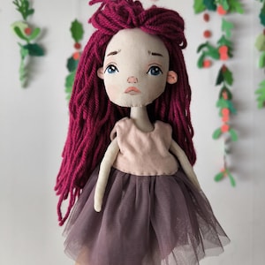 May include: Handmade cloth doll with a pale face, blue eyes, and magenta yarn hair. The doll wears a pink top and a dark purple tulle skirt. The doll's arms and legs are made of a cream-coloured fabric.