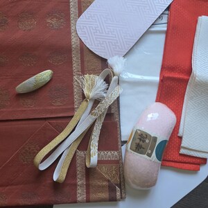 May include: A set of Japanese kimono accessories including a rust-colored obi sash with gold circular patterns, a white patterned obi-ita board, a pink obi-makura cushion, a red fabric strip, a white textured sash, and a gold and white hair clip.