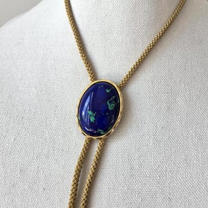 May include: A gold-toned bolo tie featuring an oval-shaped, deep blue stone with green inclusions. The stone is set in a gold frame and hangs from a woven gold cord, displayed on a mannequin.
