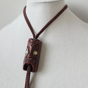 Peut inclure: Une cravate bolo marron avec un pendentif en bois sculpté en forme de visage stylisé. Le pendentif présente des incrustations irisées et vertes. Le cordon tressé est marron et le bijou est présenté sur un mannequin blanc.