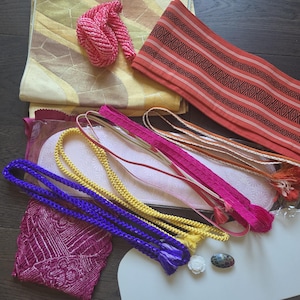 May include: A collection of Japanese kimono accessories including a folded yellow and brown patterned fabric, a red sash with black geometric designs, several braided cords in purple, yellow, and pink, a small white flower ornament, and a dark stone.