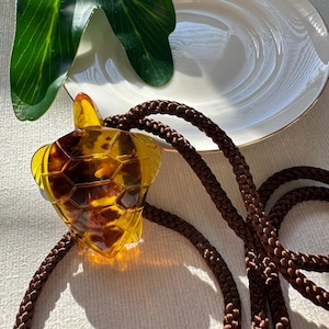 May include: A turtle-shaped pendant with a brown and yellow tortoiseshell design, suspended from a brown braided cord. The pendant is made of a translucent material. The cord is knotted. The background includes a white plate with a gold rim and green leaves.