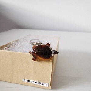 May include: A small, brown turtle keychain with a silver chain. The keychain is in a brown box with the text "turtosie11pin" on the front.