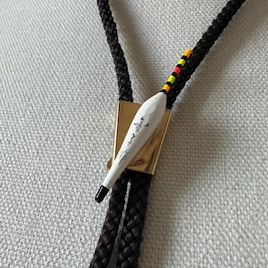 Peut inclure: Une cravate bolo avec un cordon tressé marron foncé. La cravate présente un pendentif blanc en forme de goutte d'eau avec du texte noir et une pointe noire. Le pendentif est orné de rayures jaunes, rouges et orange.