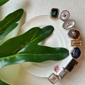 May include: A collection of decorative brooches and pins in various shapes and designs, including square, oval, and rectangular. The pins feature gold, silver, and enamel finishes, with some displaying the Burberry logo. The items are arranged on a white plate with a gold rim.