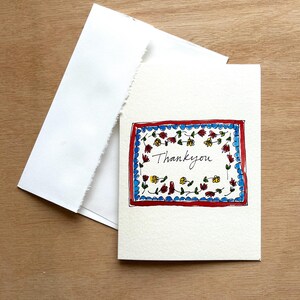 May include: A white card with a red border and a blue and white floral design inside. The text "Thank you" is written in black ink inside the floral design.