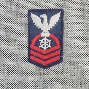 May include: A rectangular embroidered patch featuring a white eagle perched above a white ship's wheel centered within a red circular border, set over two red horizontal chevrons on a dark blue background, attached to a textured gray fabric.