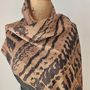 May include: A tan and black patterned scarf draped over a black dress form. The scarf features a bold, abstract design in shades of tan and black, creating a striking visual contrast. The scarf's fabric appears to be lightweight and flowing.