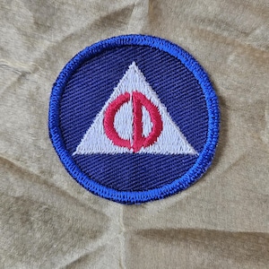 May include: A circular patch with a blue border and a white triangle at the center. Inside the triangle is a red symbol. The patch is sewn onto a tan fabric.
