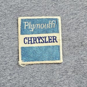 May include: A rectangular embroidered patch featuring a blue background with the white text Plymouth on the top section and the blue text CHRYSLER on a white horizontal center stripe, all bordered by a cream-colored stitched edge on gray fabric.