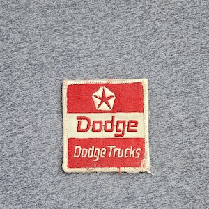 Puede incluir: Un parche rectangular bordado con un diseño en rojo y blanco que incluye un emblema de estrella central. El texto dice Dodge en letras rojas grandes y Dodge Trucks en letras blancas más pequeñas sobre una tela de color azul jaspeado.