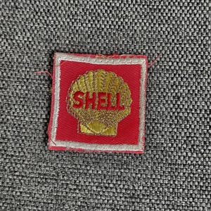 Peut inclure: Un écusson brodé carré présentant un fond rouge avec une bordure blanche. Au centre, un motif de coquille Saint-Jacques dorée est brodé avec le mot SHELL en lettres capitales rouges au milieu du graphisme de la coquille.