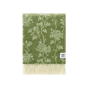 May include: A rectangular olive green throw blanket featuring a white floral rose pattern across the surface. The blanket has a white fringe along the bottom edge and a small white rectangular fabric tag with the black text HOME LIKES.