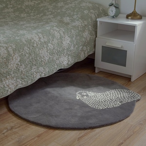 Op de afbeelding: Een rond grijs tapijt met een wit schaapontwerp. Het tapijt ligt op een houten vloer naast een wit nachtkastje en een bed met een groene, bloemige bedsprei.