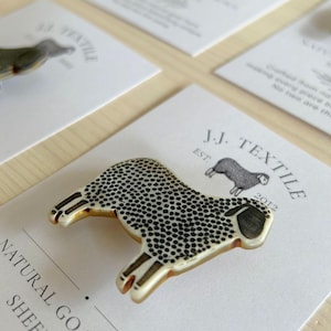 May include: A sheep-shaped brooch featuring a cream-coloured base with a detailed black dotted pattern across its body. The brooch is displayed on a white card printed with the text J.J. TEXTILE EST. 2012 and a small graphic of a sheep.