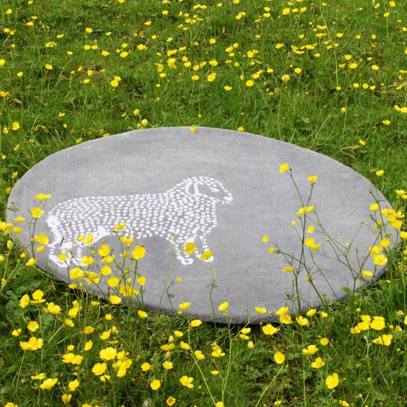 May include: A round, gray rug with a white sheep design. The rug is set in a field of green grass and yellow wildflowers. The rug is a decorative item.