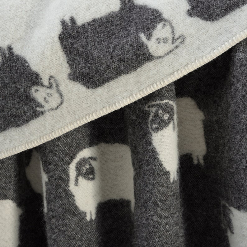 May include: A white and grey wool throw blanket with a repeating pattern of sheep and other animals. The blanket is soft and cosy, perfect for snuggling up on the sofa.