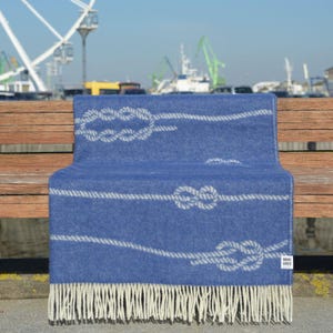 May include: A blue wool throw blanket with a white rope and knot pattern. The blanket has fringe on the bottom edge and a small white label with the text "HOME LINES" in the bottom right corner.