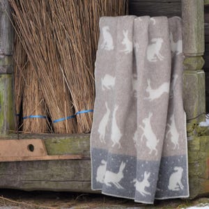 May include: A folded beige blanket with a repeating pattern of white rabbits. The blanket has a dark blue border with more rabbit silhouettes. The blanket is draped over a wooden structure with bundles of dried reeds.