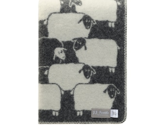 May include: A folded wool blanket featuring a repeating pattern of white sheep with black faces and legs against a dark charcoal gray background. A small gray fabric label in the corner displays the text J.J. Textile with a sheep icon.